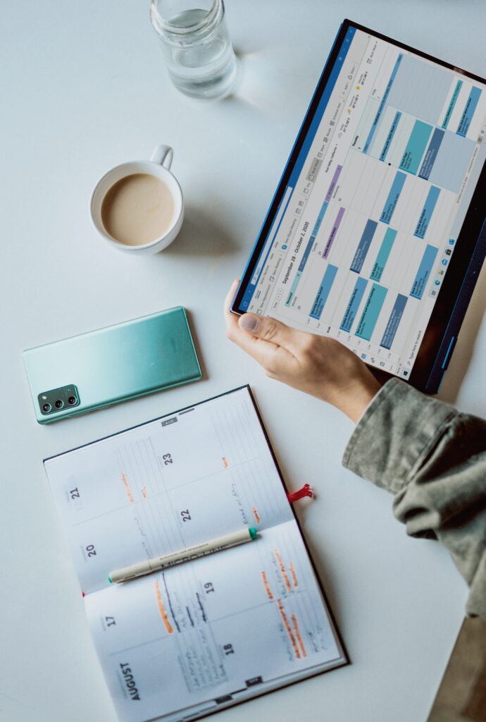 Weekly planner laid open with a pen, phone, coffee, water. While a woman looks through her calendar on her tablet. 